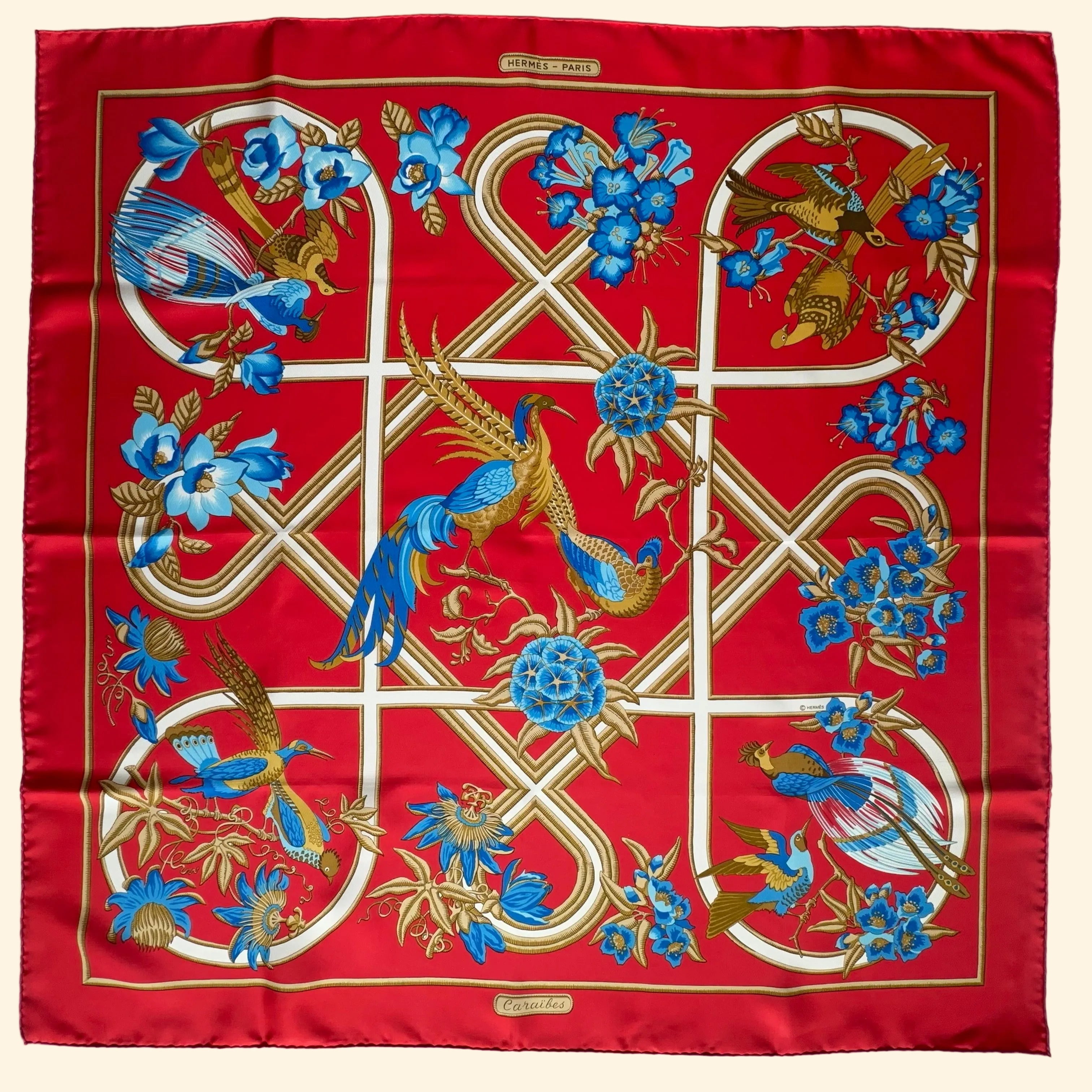 Vue d'ensemble du Carré Hermès "Caraïbes" sur fond rouge vif. Motif symétrique d'oiseaux exotiques et fleurs bleues dans un treillage blanc.